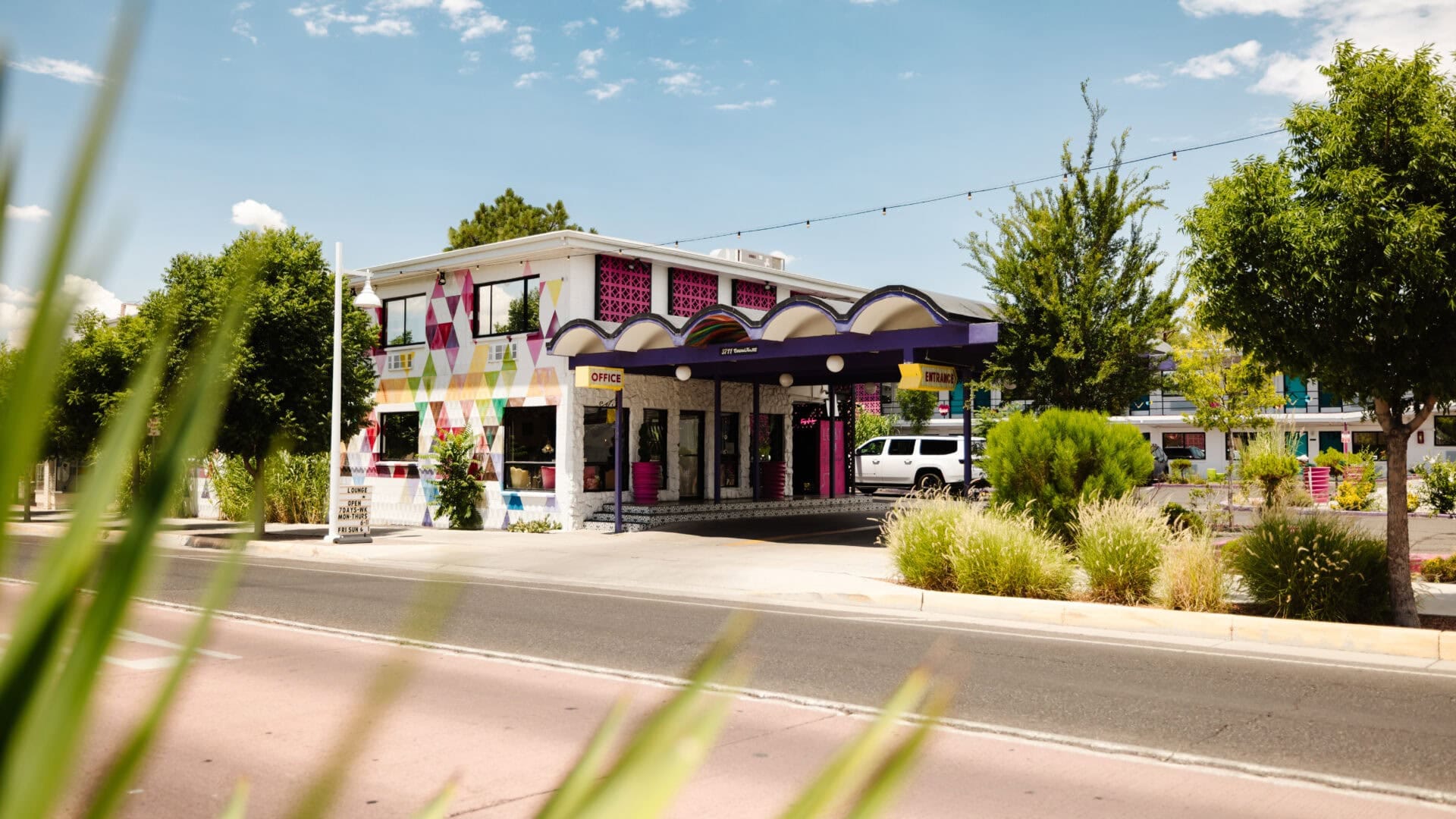 Zweistöckiges Straßenhotel mit bordeauxroten Verzierungen und überdachtem Eingang entlang Route 66 in Albuquerque, New Mexiko unter blauem Himmel mit hohem Gras im Vordergrund.
